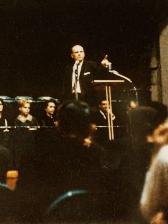 A man speaking at a podium, gesturing, with an audience seated before him in a dim setting.
